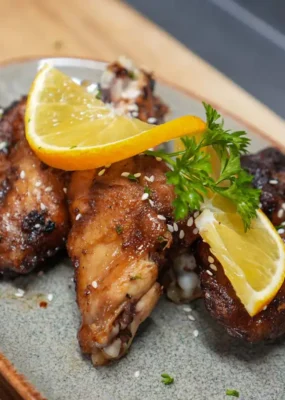 A close-up view of sticky grilled chicken wings garnished with sesame seeds, fresh parsley, and citrus wedges, served on a rustic plate at a restaurant in Lovina, Bali.